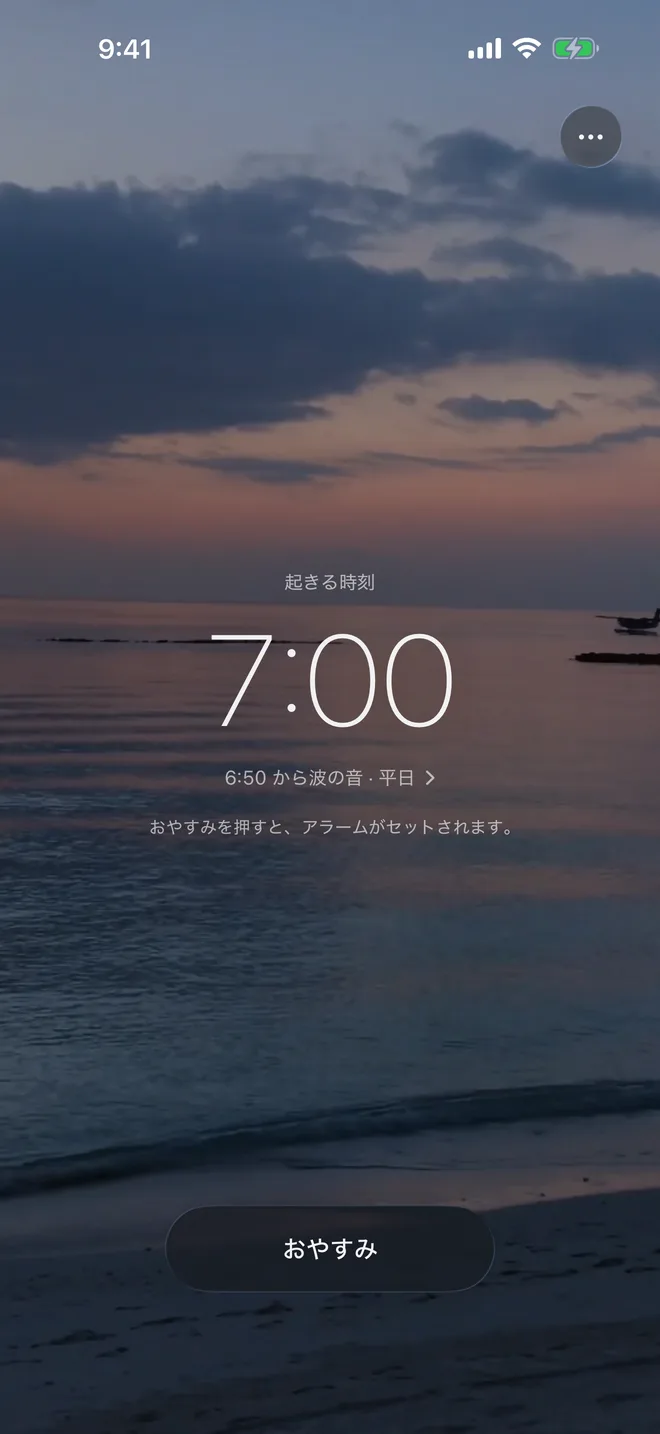 iPhone の Petit Ocean の最初の画面。夕暮れの浜に、起きる時刻 7:00 が細く大きな数字で出ている。下に「6:50 から波の音・平日」と、おやすみのボタン。